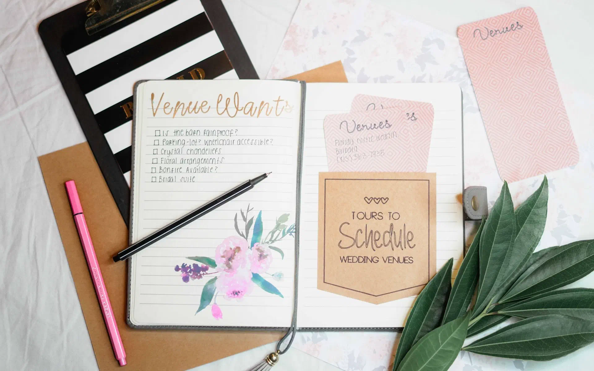 Caderno de planejamento de casamento aberto, com a lista do local dos sonhos e as visitas a agendar, entre canetas e folhas verdes